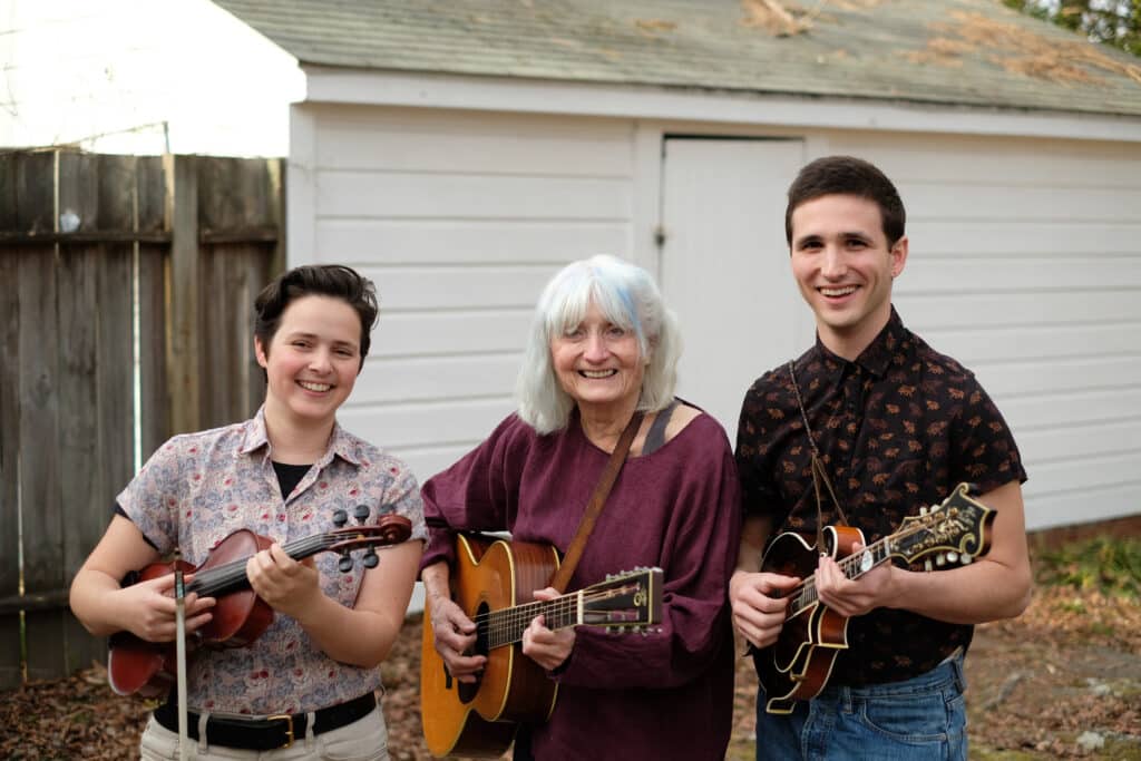 Alice Gerrard in the middle holding a guitar, Tatiana Hargreaves with fiddle and Reed Stutz holding a mandolin. They are outside in front of a white building